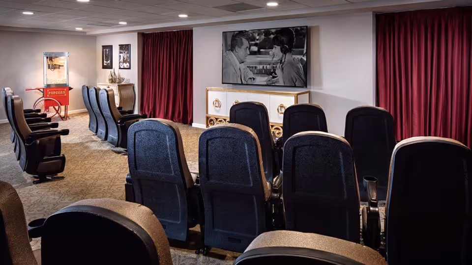 A small theater room with two rows of black cushioned seats facing a large flat-screen TV mounted on the wall. The TV displays a black and white movie scene of a man and woman talking. To the left, there is a vintage-style popcorn machine on a red cart. The room has beige carpet, white walls, and red curtains on the right side.