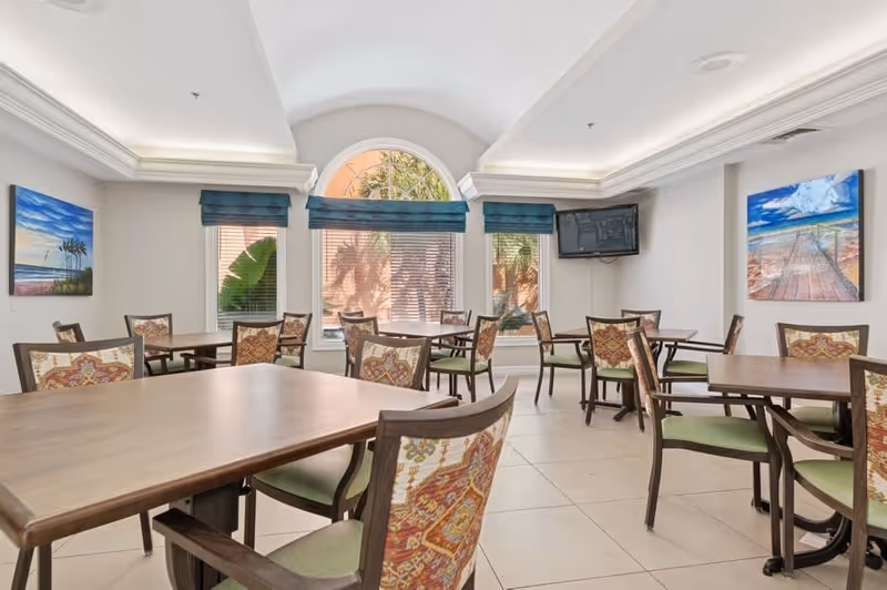 Bright dining room with multiple wooden tables and upholstered chairs, wall art, a mounted TV, and arched windows.