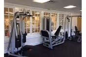 Indoor fitness room with exercise machines including a multi-station weight machine and a leg press. The room has white walls with large windows and a black carpeted floor.