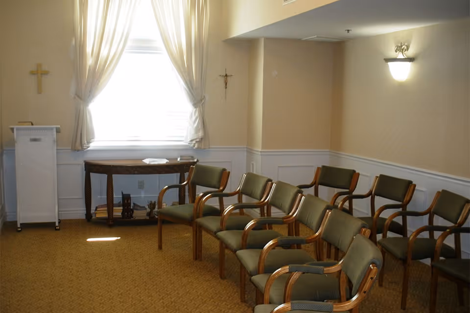 A small room with a row of green cushioned chairs with wooden armrests arranged facing a white podium and a wooden table under a window with white curtains. Two crosses hang on the walls, one on each side of the window. The room has beige walls with white wainscoting and a wall-mounted light fixture.