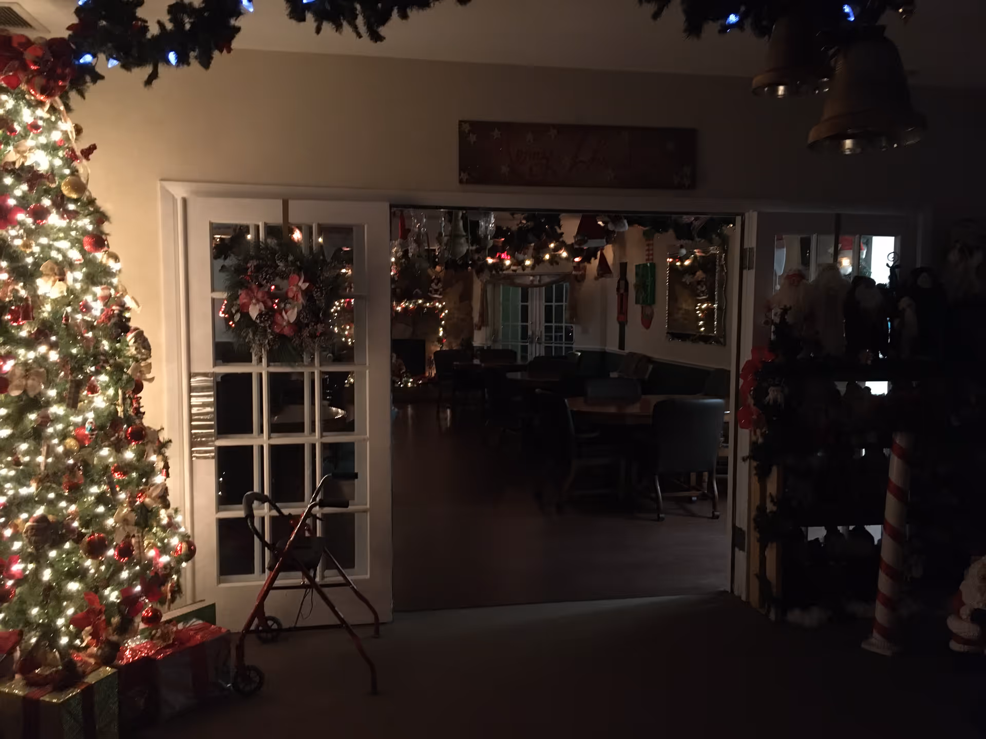 Dimly lit interior of a senior living facility decorated for Christmas with a large, brightly lit Christmas tree on the left, wrapped presents underneath, a red walker in front of glass-paneled double doors adorned with a wreath, and a dining area visible through the doors with tables and chairs. Garland and holiday decorations hang from the ceiling and walls.