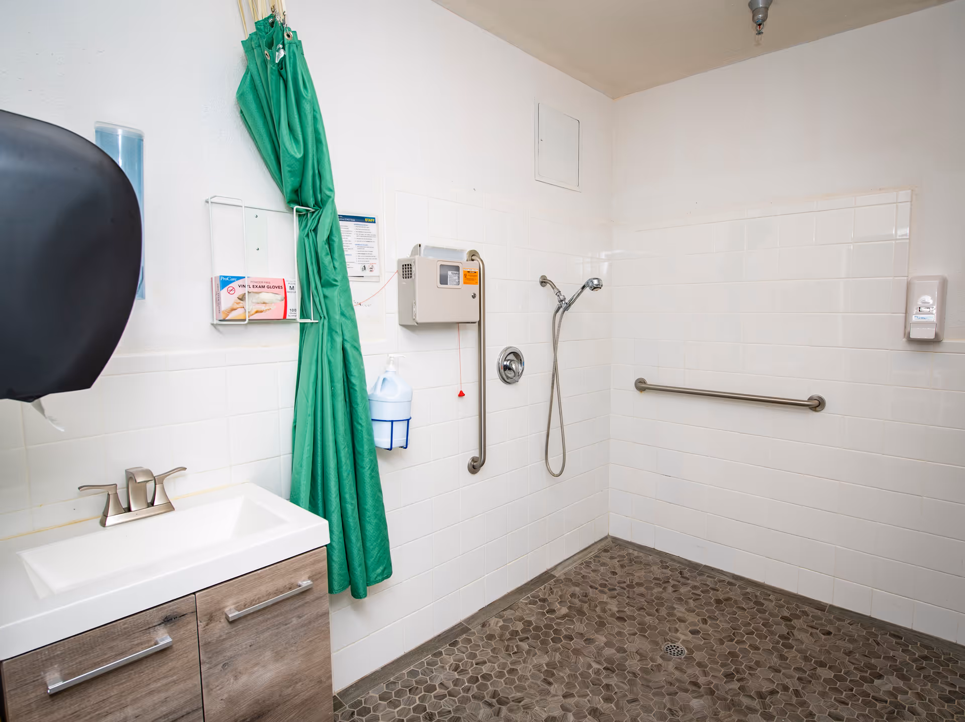 A clean, accessible shower room with white tiled walls and a pebble-textured floor. The room features a handheld showerhead, a grab bar on the wall, a green privacy curtain, a sink with a wooden cabinet, a soap dispenser, and a wall-mounted paper towel dispenser.
