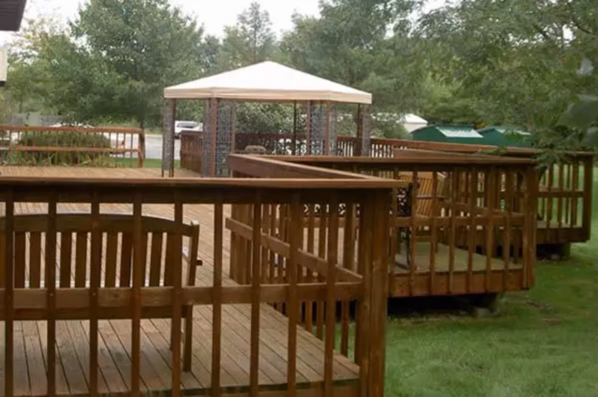 Outdoor wooden deck area with railings and benches, featuring a canopy-covered seating area surrounded by trees and greenery.