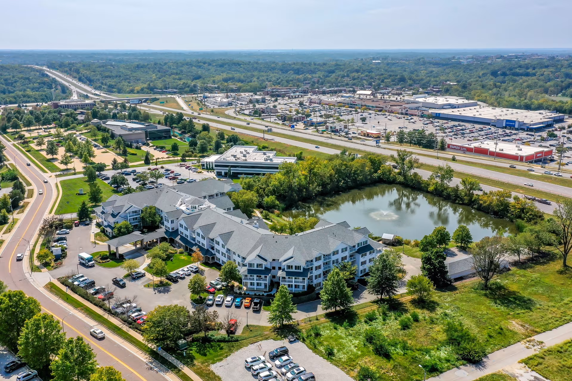 Aerial view of Solstice Senior Living at Columbia facility showing a large multi-wing building surrounded by trees, parking lots, and a pond with a fountain. Nearby roads, highways, and commercial buildings are visible in the background under a clear sky.