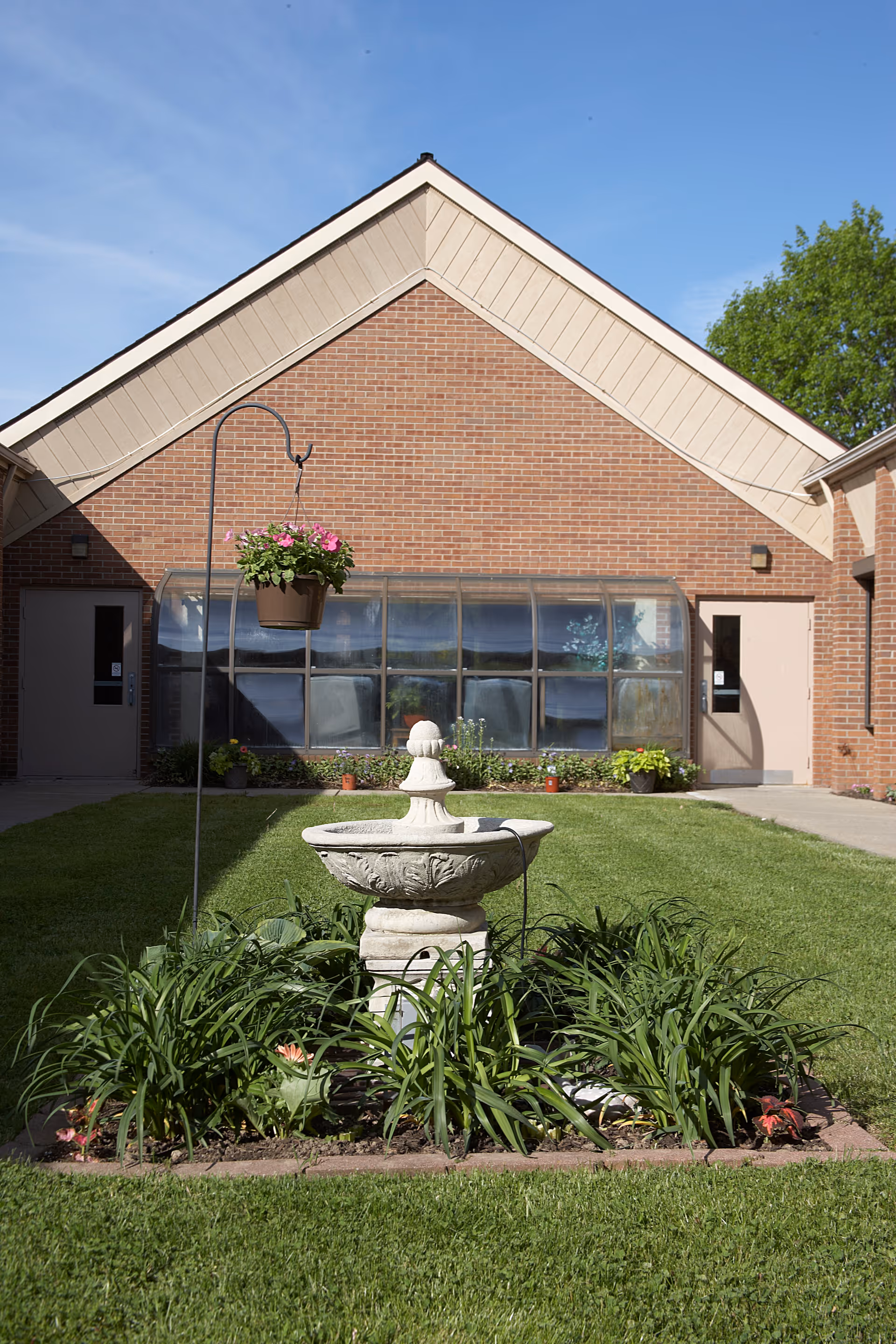 A courtyard area with a decorative stone fountain surrounded by green plants and flowers. Behind the fountain is a brick building with a large window and two doors on either side. A hanging flower pot with pink flowers is suspended on a metal hook in the foreground. The sky is clear and blue.
