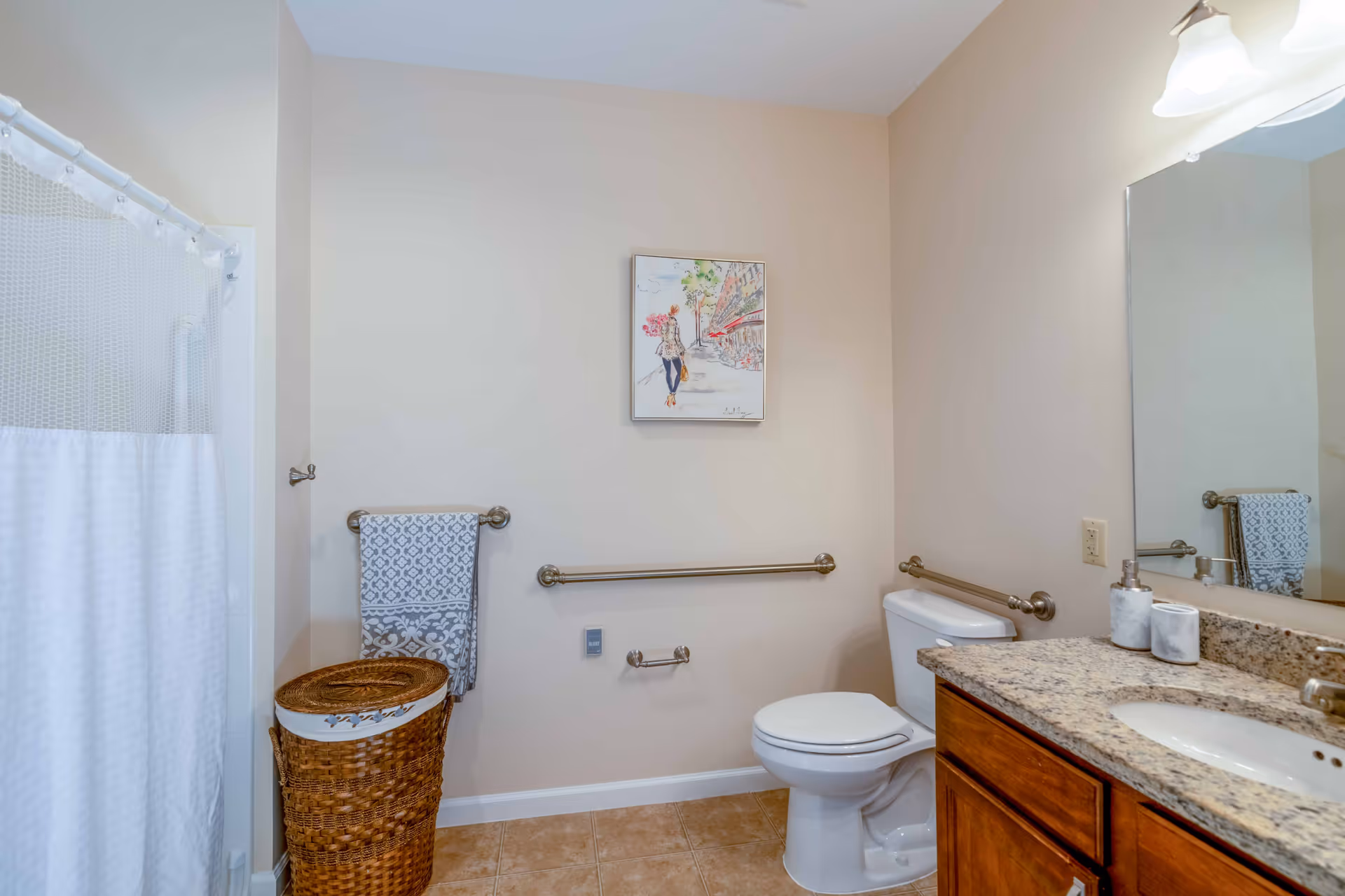 A tidy assisted-living bathroom featuring a toilet, sink with granite countertop, shower with curtain, grab bars, towel rack, and a wicker laundry basket.