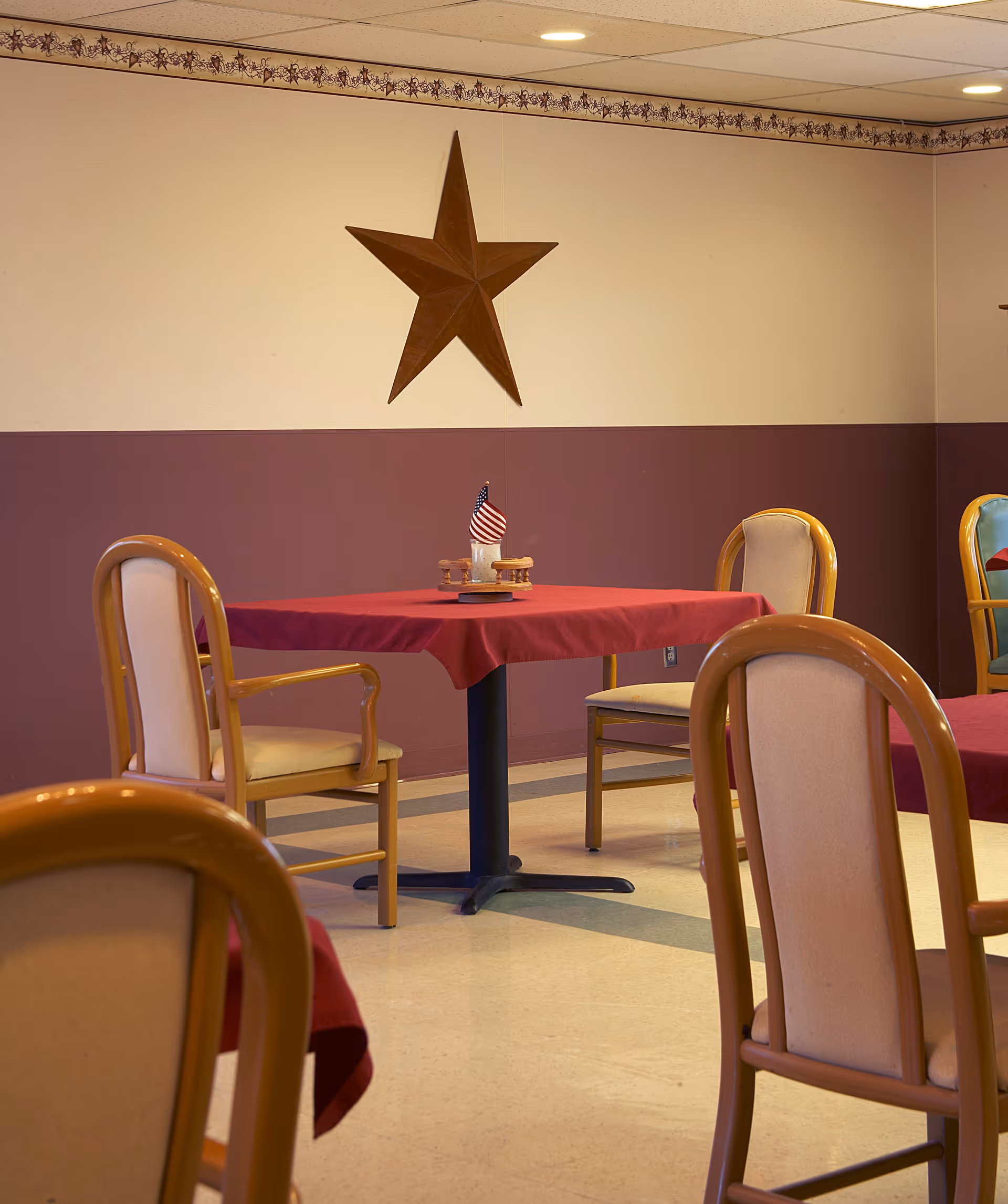 A dining area with tables covered in red tablecloths and wooden chairs with beige cushions. A small centerpiece with an American flag is on one table. The walls are painted half beige and half maroon with a decorative border near the ceiling and a large wooden star mounted on the wall.