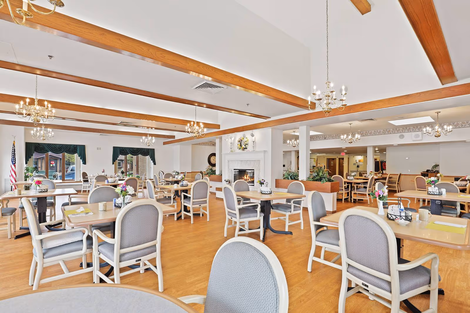 Spacious senior living dining room with multiple tables and chairs, chandeliers, a fireplace, and large windows.