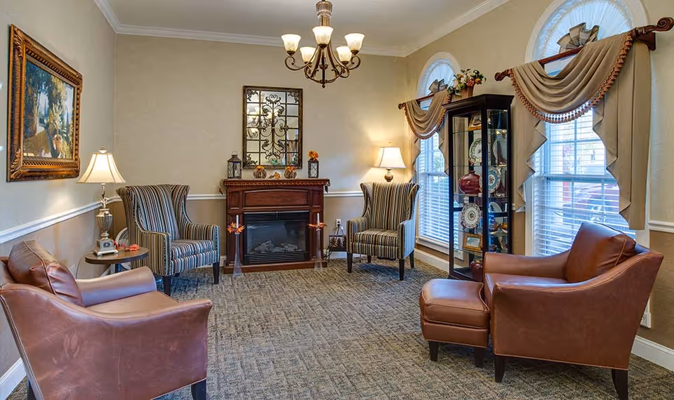 A cozy senior living room with two brown leather armchairs, two striped armchairs, a small wooden fireplace, a glass display cabinet with decorative plates, two large windows with draped curtains, a chandelier, a table lamp, and a framed painting on the wall.