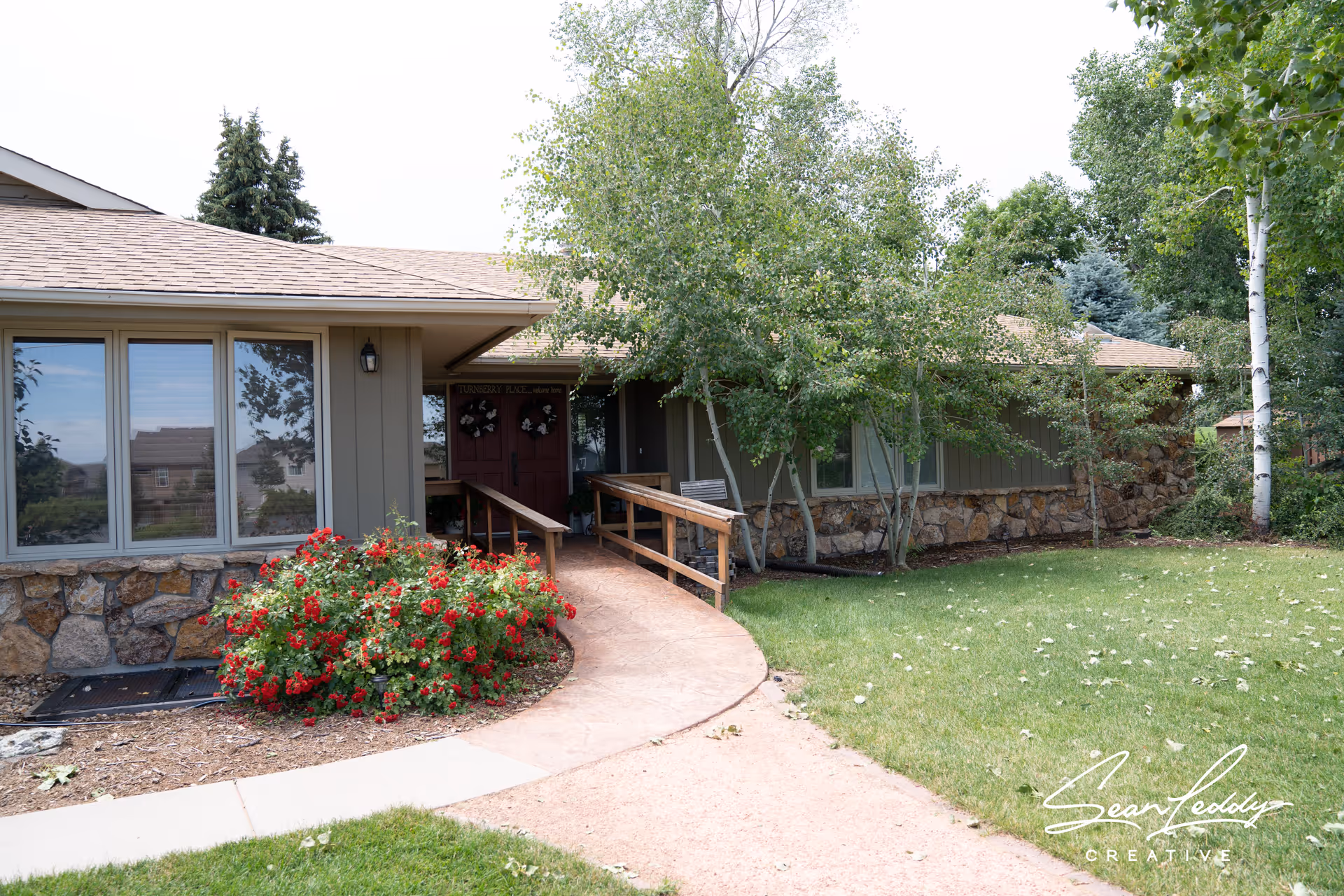Single-story assisted living building entrance with a ramp, stone facade, flowerbed and surrounding trees.