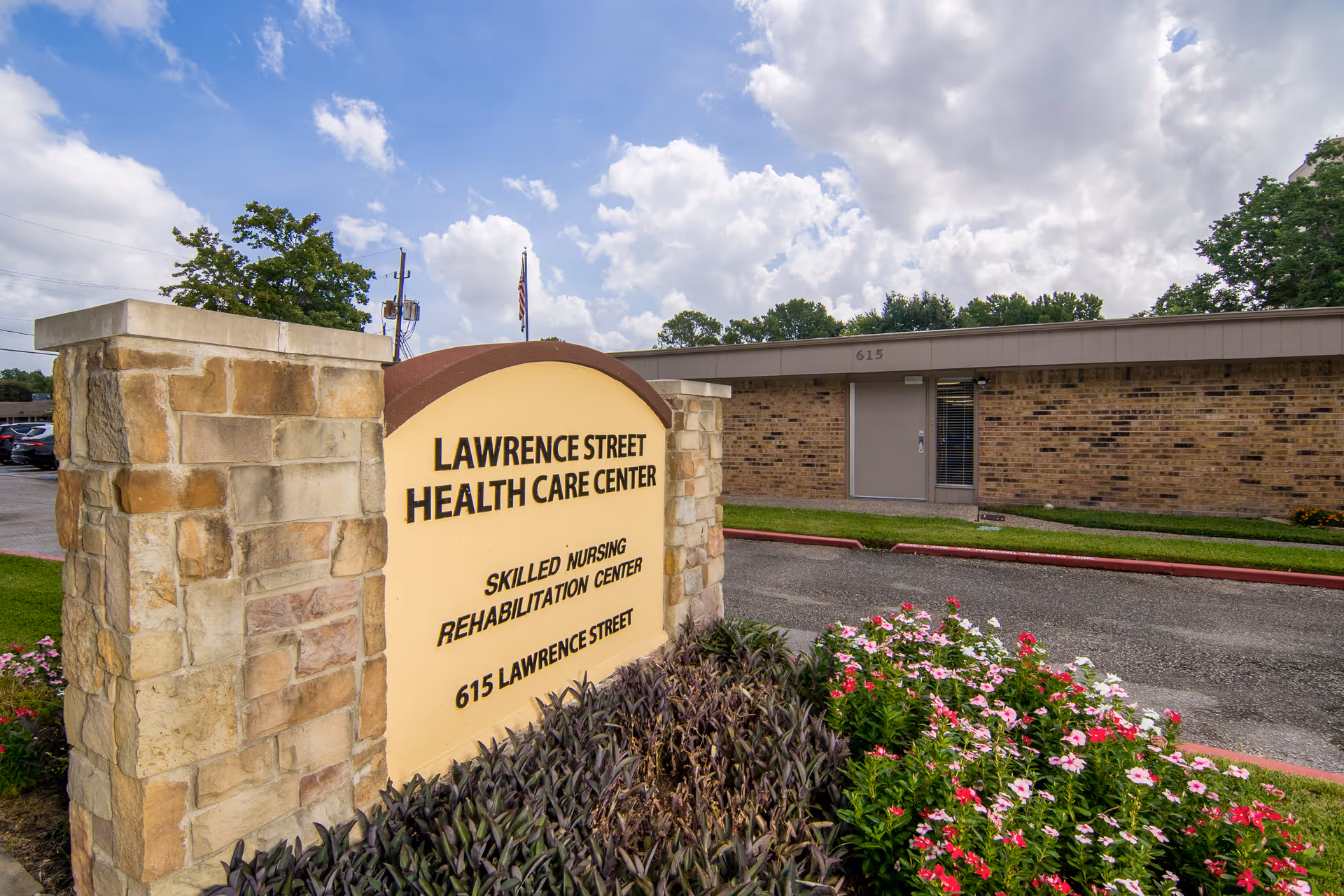 Stone entrance sign reading 'Lawrence Street Health Care Center' in front of a single-story brick building with flowers and a cloudy sky.