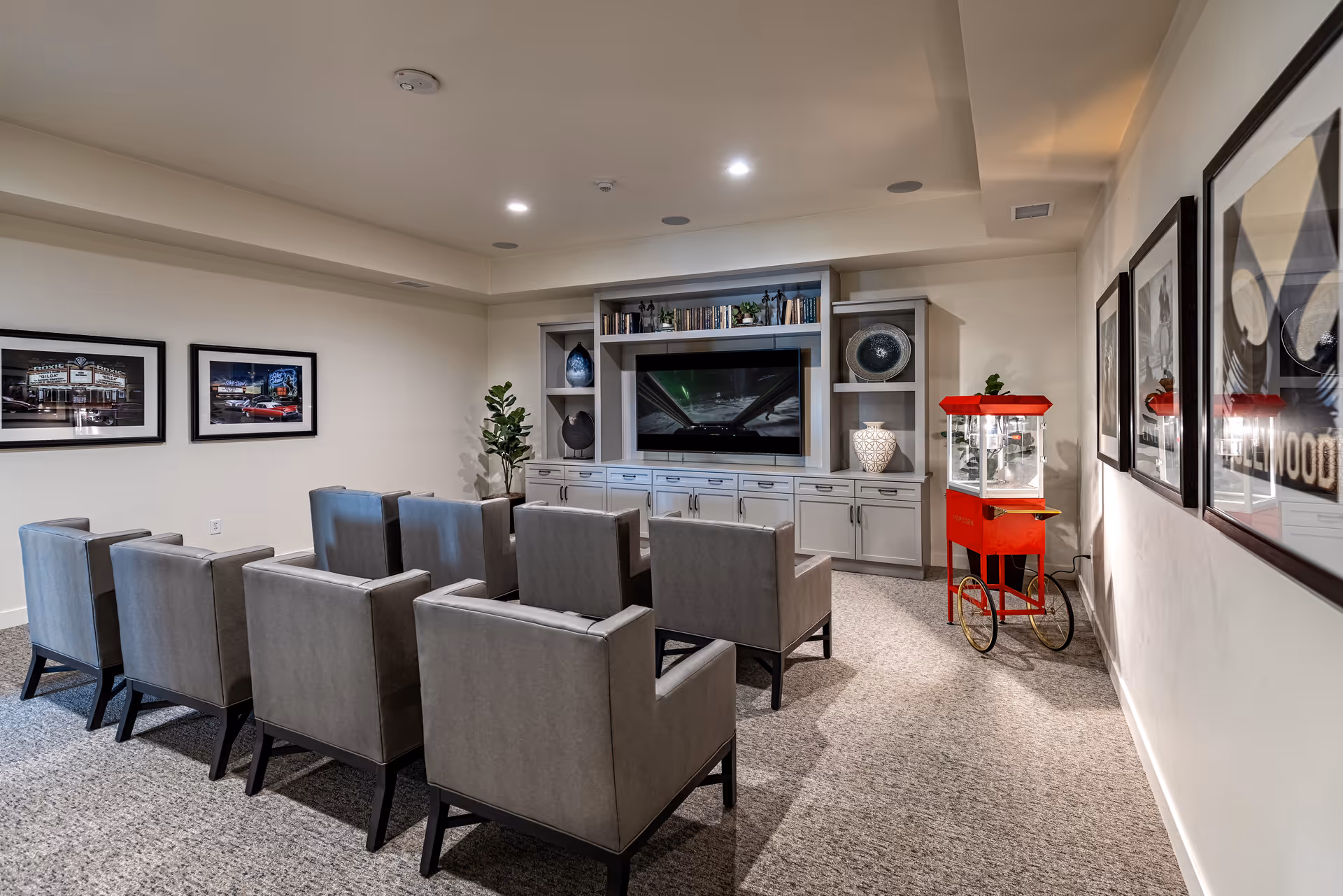 A cozy media room with two rows of gray upholstered chairs facing a large flat-screen TV mounted on a built-in cabinet with shelves displaying decorative items and books. The room has framed pictures on the walls and a red popcorn machine on wheels in the corner.