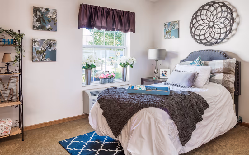 A cozy bedroom with a neatly made bed featuring white and gray bedding and multiple pillows. A decorative tray with glasses and a book rests on the bed. The room has a window with a purple valance, two flower arrangements on the windowsill, and wall art including floral paintings and a circular metal wall decoration. A small nightstand with a lamp and a photo frame is beside the bed, and a metal shelving unit with decorative items is on the left side of the room.
