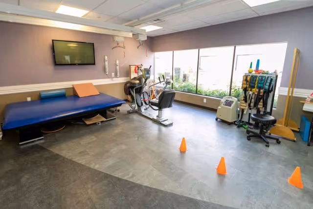 A bright rehabilitation room with a treatment table, stationary bike, therapy equipment, orange cones, a wall-mounted TV, and large windows.
