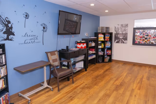 A cozy room with wooden flooring and light blue and white walls. There is a wall-mounted TV above a black console table with various items on it, including a red box. Next to the table is a wooden chair with a cushion and a small rolling table. On the blue wall, there is a decorative decal featuring a fairy, dandelions, and the phrase 'a dream is a wish your heart makes.' Two black shelving units hold board games and other items. On the white wall, there are two framed pictures and a bulletin board filled with photos and notes.
