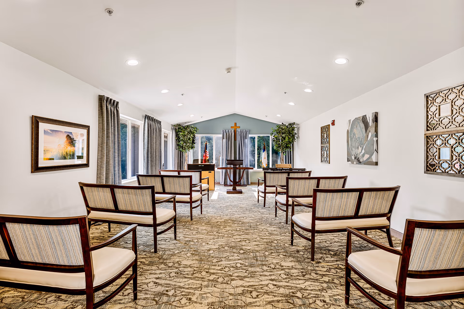 A bright, well-lit room arranged for a small gathering or service with rows of wooden chairs with cushioned seats facing a podium and a wooden cross on the far wall. The room has large windows with curtains, framed artwork on the walls, and two potted plants near the front.