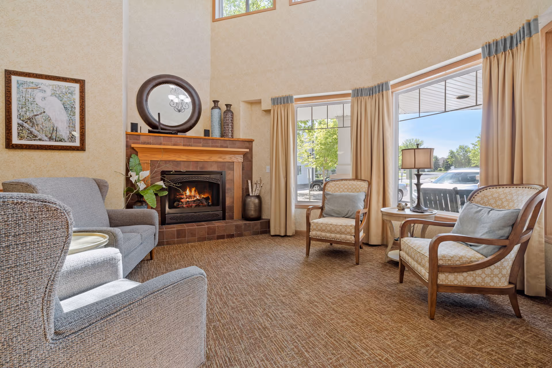 A cozy living room area with a lit fireplace surrounded by brown tiles and a wooden mantel. Above the mantel is a round mirror and decorative vases. The room has beige patterned wallpaper and large windows with beige curtains letting in natural light. There are two upholstered armchairs with wooden frames and blue cushions, a small round wooden side table with a lamp, and a gray sofa and armchair. A framed picture of a bird hangs on the wall to the left.