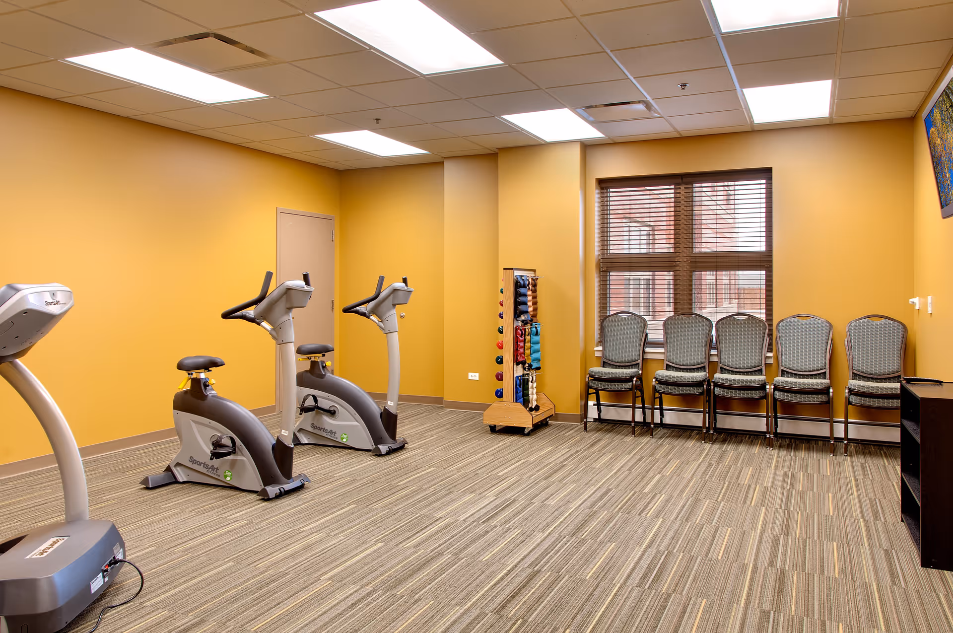 A fitness room with two stationary exercise bikes, a rack of colorful hand weights, and a row of five chairs lined up against a wall with a window. The walls are painted yellow, and the floor is carpeted with a striped pattern. The ceiling has recessed lighting panels.