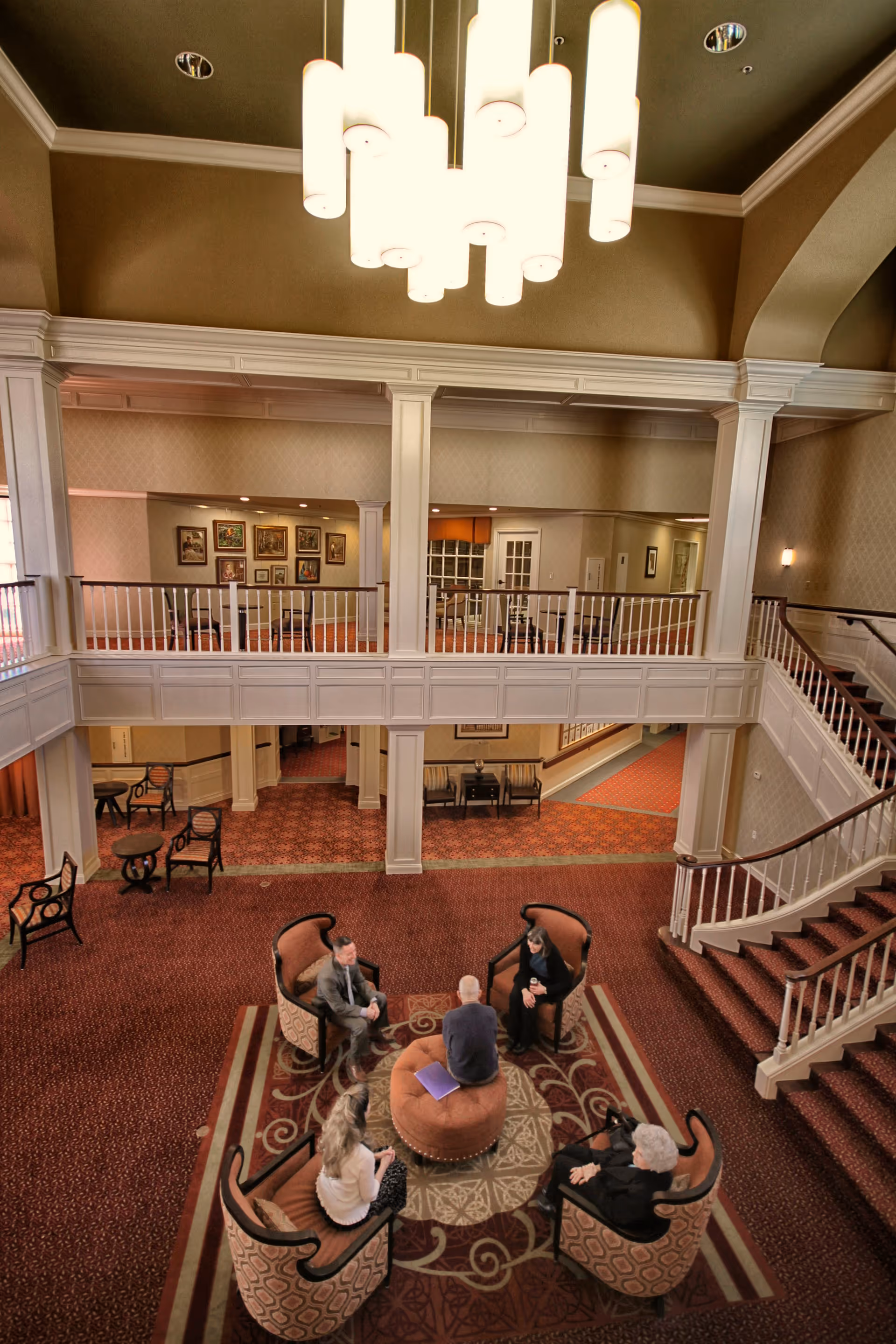 A spacious, elegant common area in a senior living facility with five people seated in a circle on patterned armchairs around a round ottoman. The room features a red carpet with intricate designs, white railings on the upper level, and a large modern chandelier hanging from the ceiling. A staircase with white banisters leads to the upper floor, and framed artwork decorates the walls.