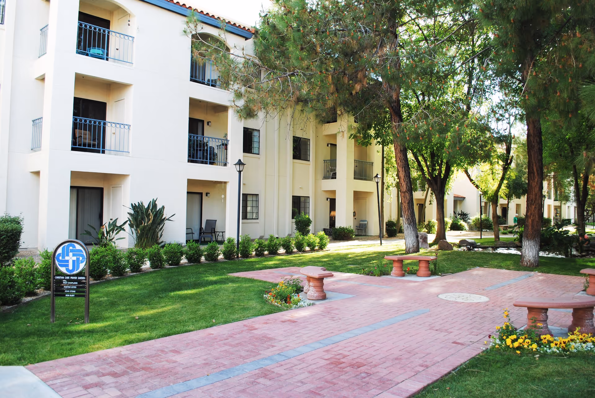 Outdoor view of a senior living facility with a three-story beige building featuring balconies and patios. The foreground shows a paved walkway with benches and landscaped greenery including trees, bushes, and flowers.