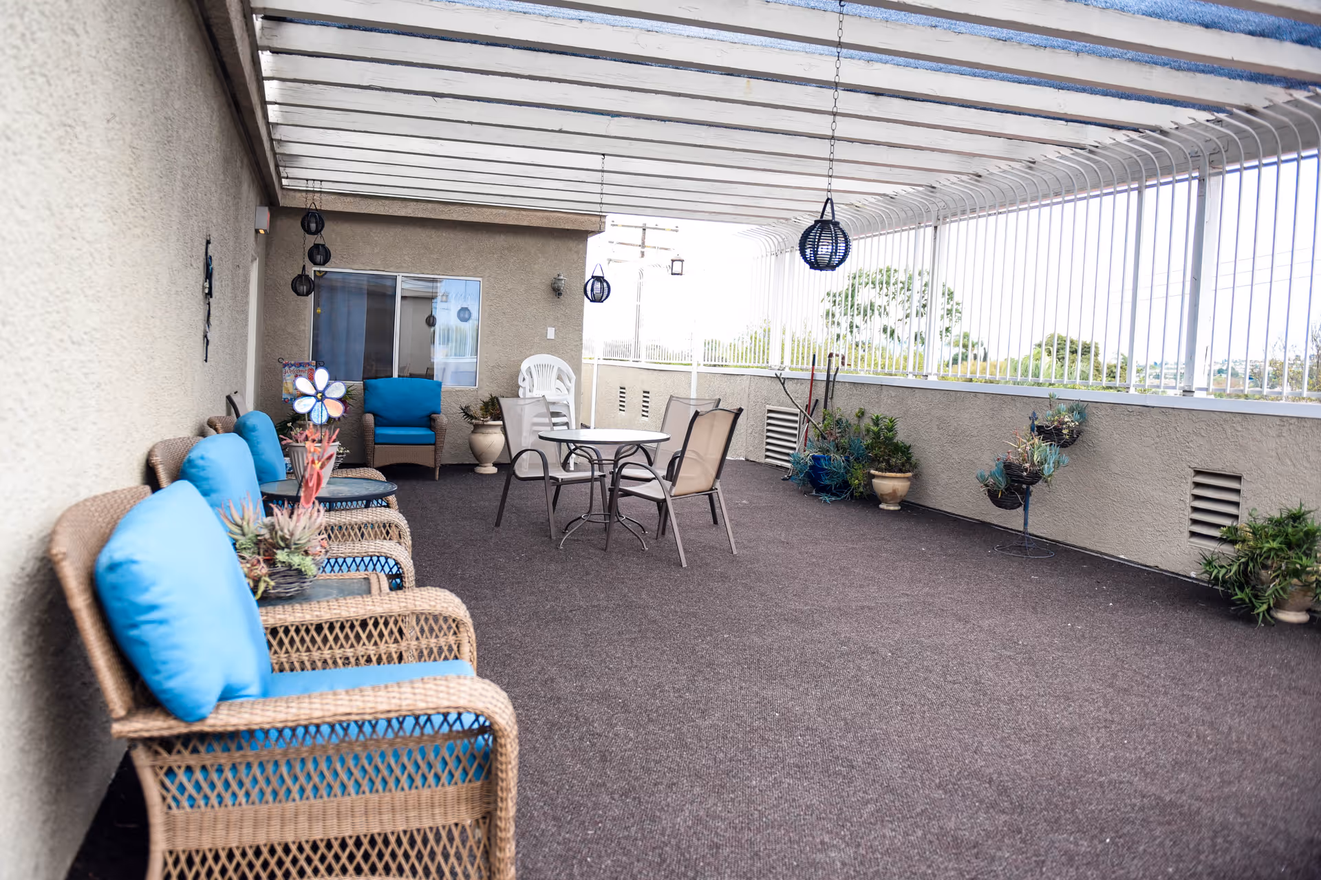 Covered outdoor patio area with wicker chairs featuring blue cushions, a small round table with chairs, hanging lanterns, and various potted plants along the walls and floor.