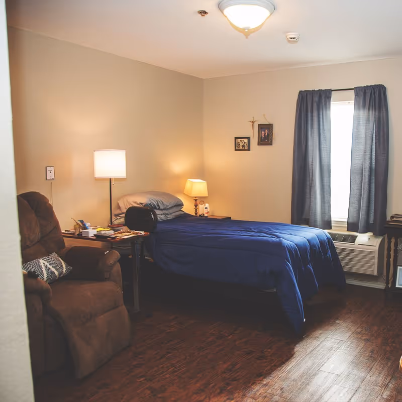 A cozy senior living bedroom with a single bed covered in a blue comforter, a brown recliner chair with a patterned pillow, two bedside lamps, framed photos and a crucifix on the wall, a window with dark curtains, and a wooden floor.