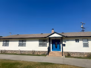 Single-story light-colored building with a blue front door, steps, and a small lawn under a clear blue sky.