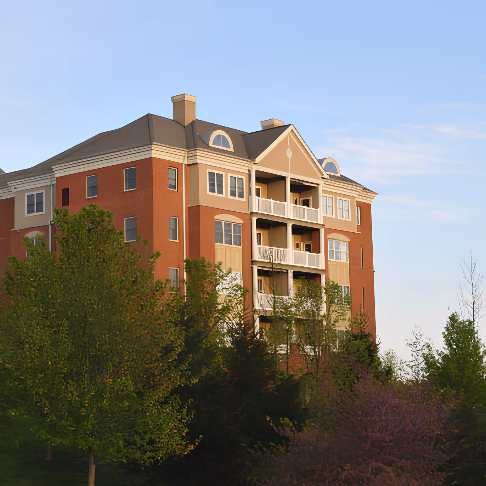 Multi-story red-brick residential building with balconies and white trim surrounded by trees under a clear blue sky.