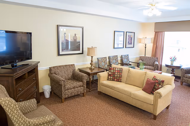 A cozy living room in an assisted living facility featuring a beige sofa with patterned throw pillows, several patterned armchairs, a wooden TV stand with a flat-screen television, framed artwork on the walls, a side table with a lamp, and large windows with curtains allowing natural light to fill the room.