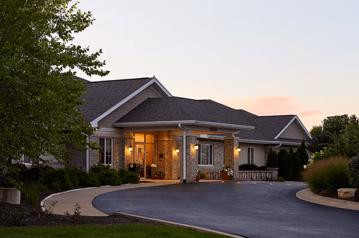 Exterior view of a single-story senior living facility building at dusk with a covered entrance, warm lights glowing, surrounded by greenery and a curved driveway.