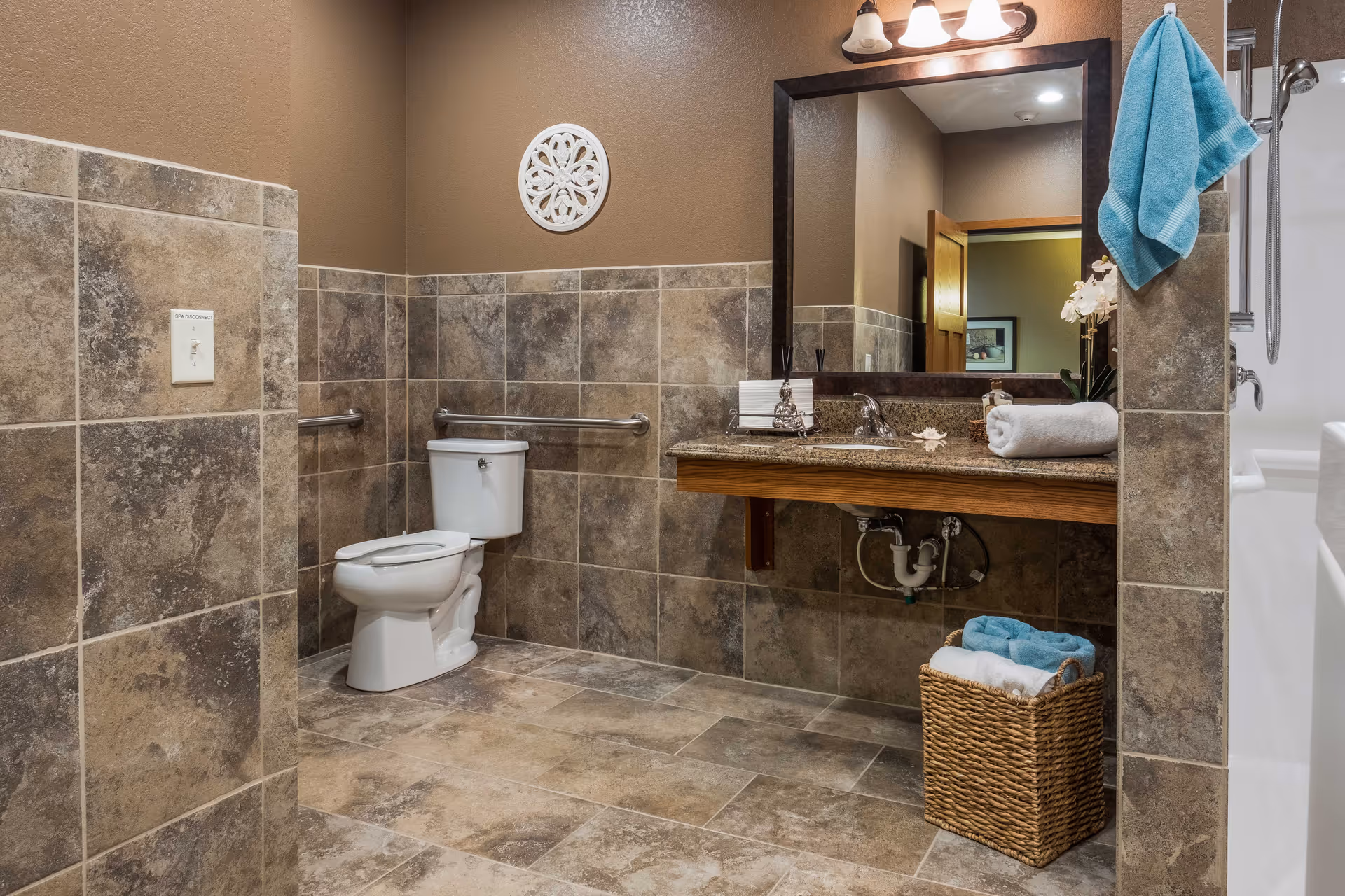 A spacious bathroom with brown tiled walls and floor, featuring a white toilet with grab bars on the walls, a large mirror above a granite countertop with a sink, a rolled white towel on the counter, a wicker basket with folded towels underneath, and a blue towel hanging near the shower area.