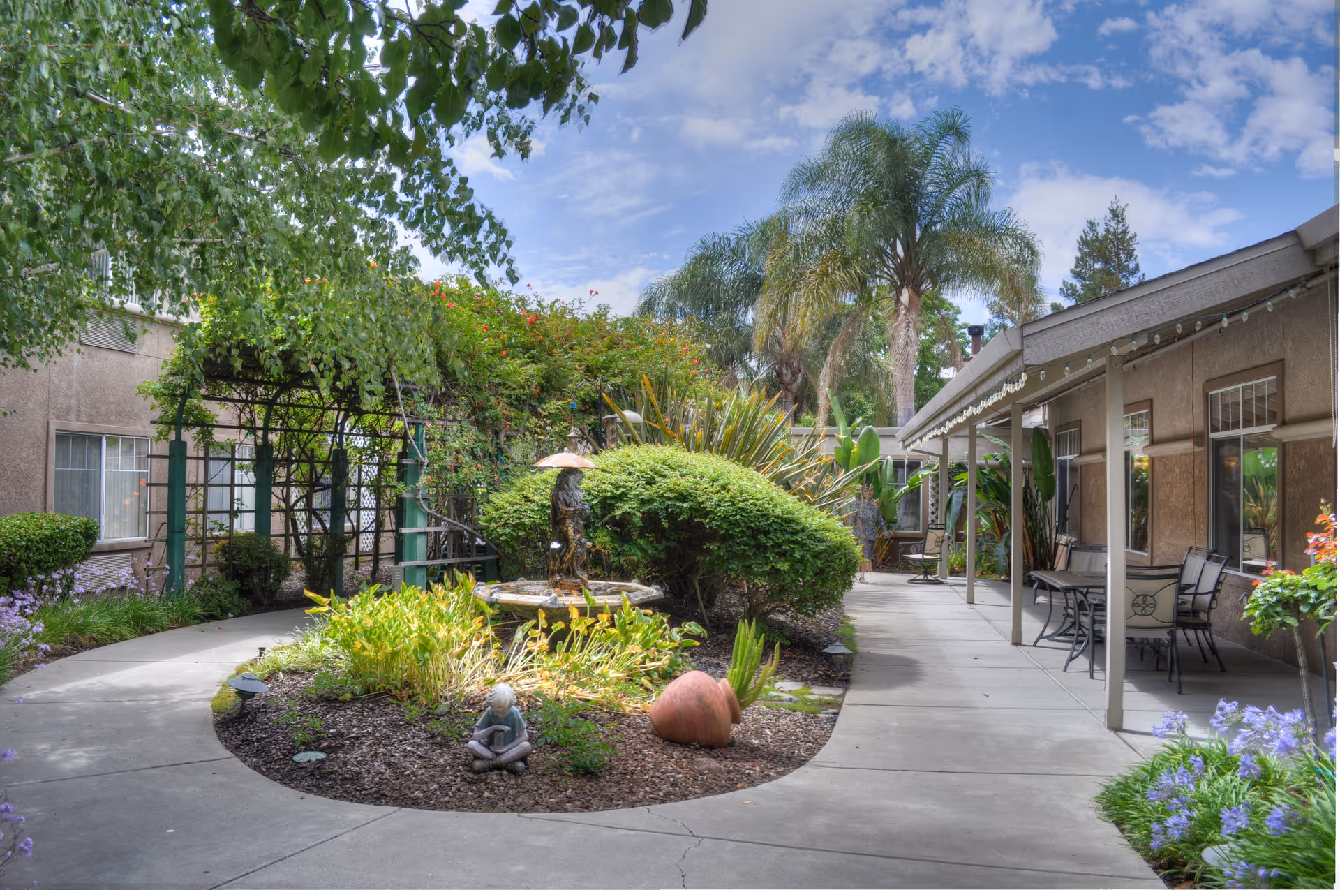 Outdoor courtyard area at Carlton Senior Living Downtown Pleasant Hill featuring a curved concrete walkway around a garden bed with various plants, a small statue of a child reading, and a fountain sculpture of a child holding an umbrella. There are palm trees and other greenery in the background, with a covered patio area on the right side furnished with tables and chairs.