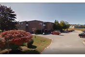 Brick three-story senior living building with a parking lot, landscaping, and a few parked cars under a clear sky.