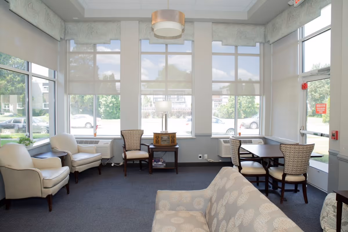 A bright and airy living room area with large windows letting in natural light. The room features two cream-colored armchairs, a patterned sofa, two wooden chairs with patterned upholstery, a small wooden table with a vintage-style radio and a lamp, and a dark wooden table with two chairs near the glass door. The room has a gray carpet and light-colored walls with window shades partially drawn.