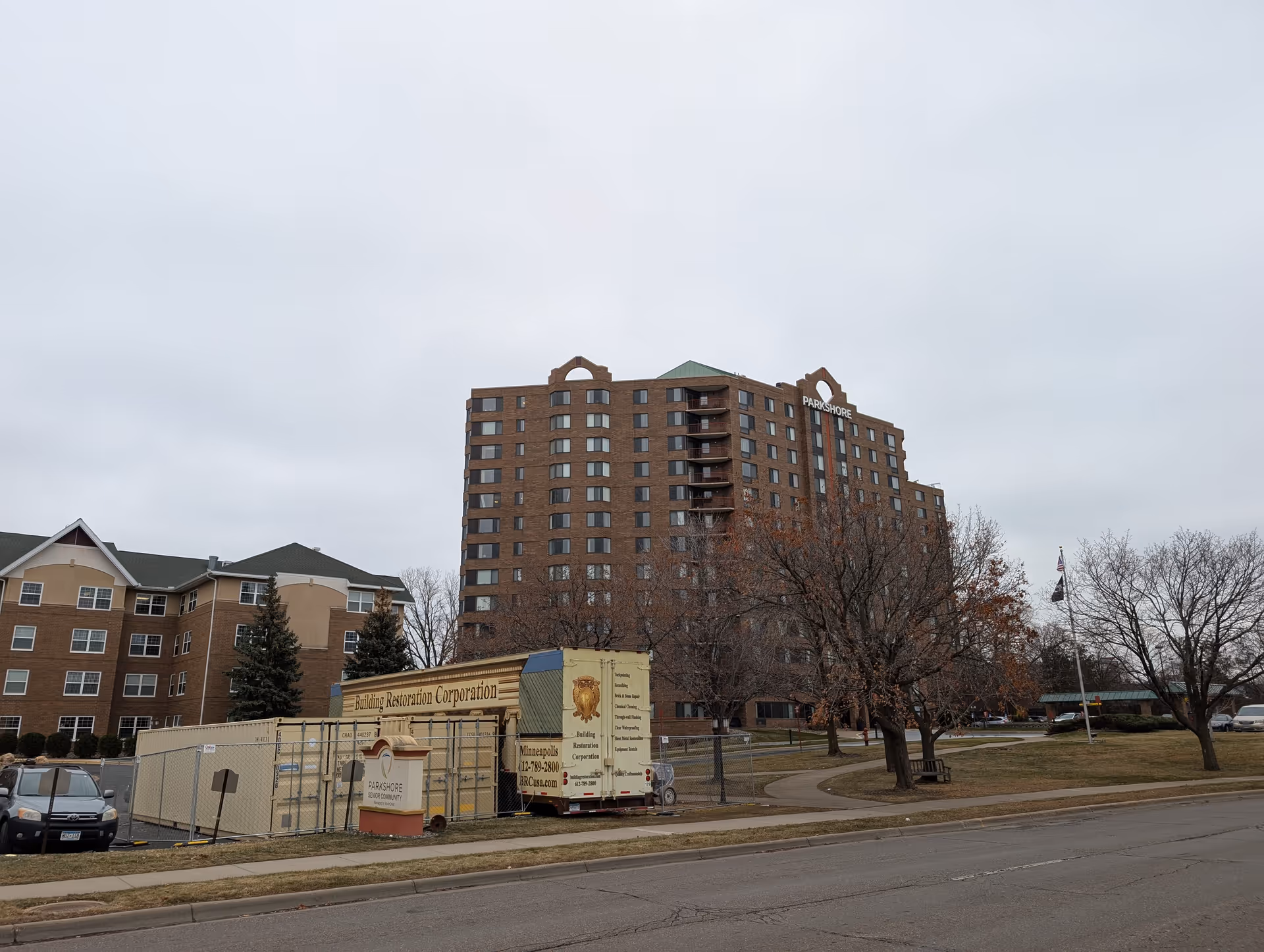 Exterior front view of a multi-story brick senior living building labeled "Parkshore" with fenced restoration trucks and lawn in front.