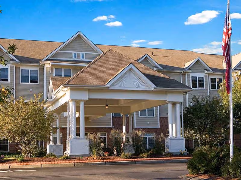 Exterior view of a senior living facility building with a covered entrance supported by white columns, surrounded by trees and landscaping, under a blue sky with scattered clouds. An American flag is visible on the right side.