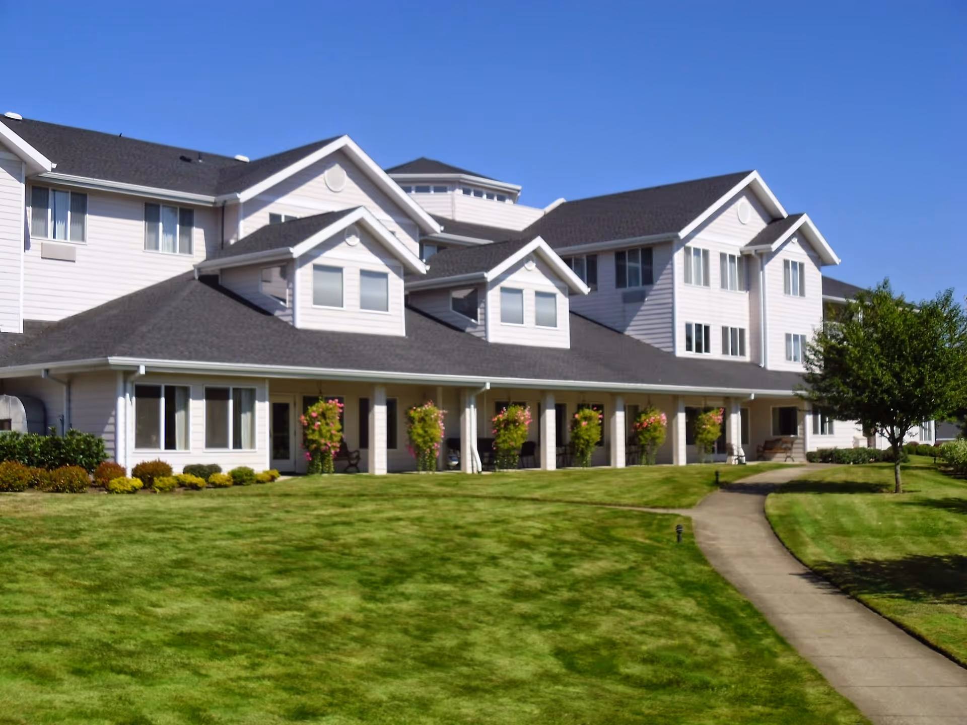Exterior view of a large, multi-story assisted living facility building with white siding and dark gray roofs under a clear blue sky. The building has multiple windows and a covered porch with hanging flower baskets. In front, there is a well-maintained green lawn with a curved concrete walkway and a small tree on the right side.