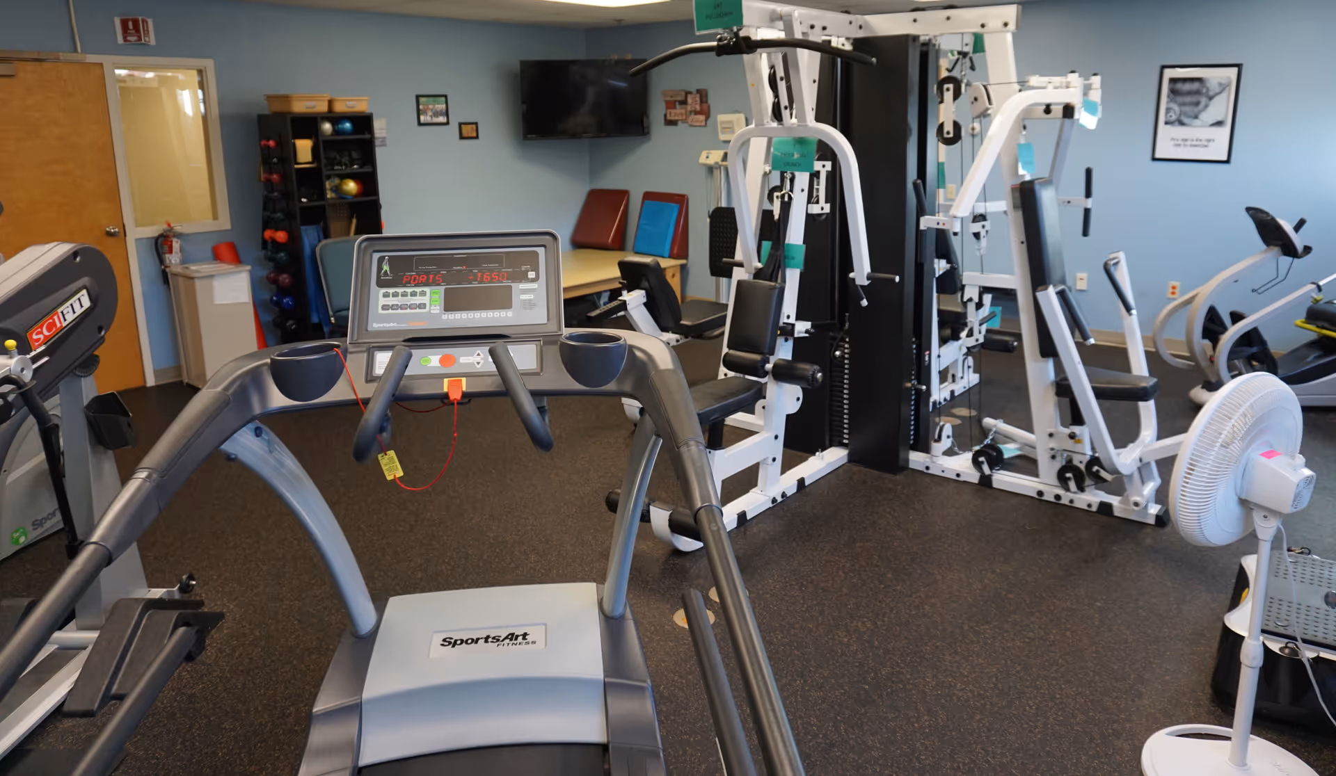 Interior view of a fitness room with various exercise equipment including a treadmill, weight machines, and stationary bikes. The room has light blue walls, a wall-mounted TV, and a rack with colorful exercise balls and weights. There is a door and a fire extinguisher visible on the left side.