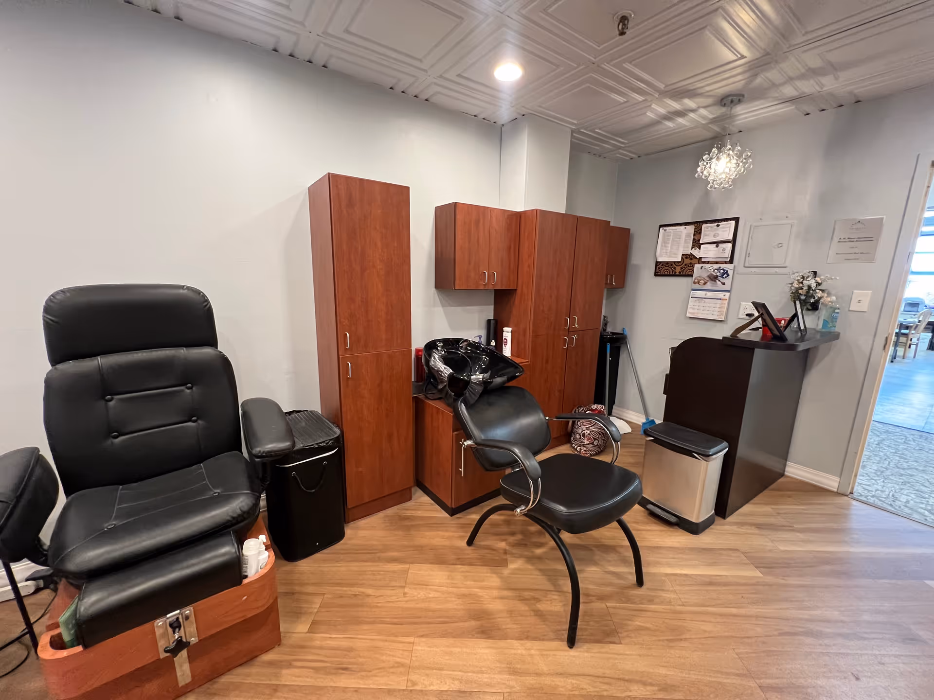 Small salon area with black salon chairs, a shampoo sink, and wooden storage cabinets on a wood floor.
