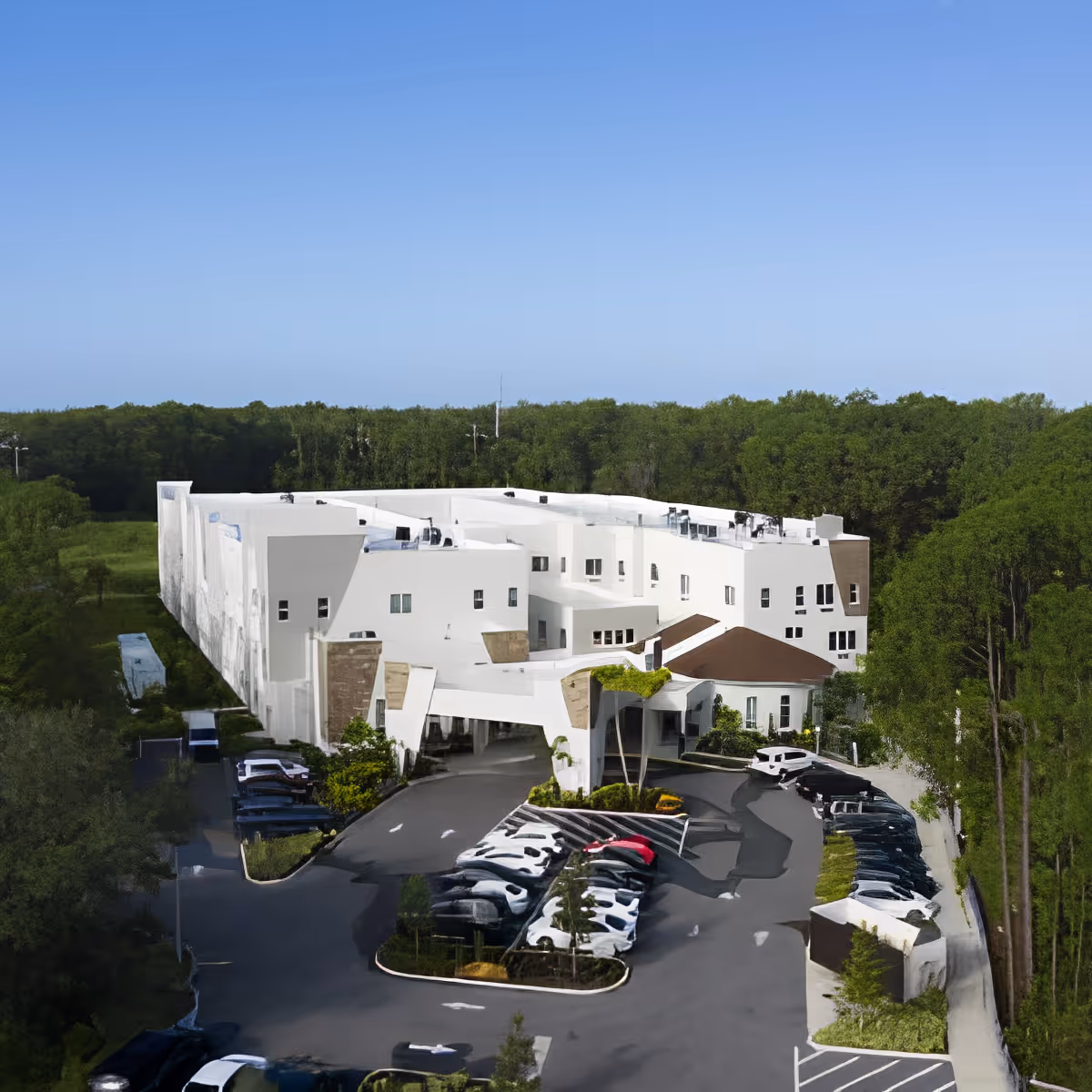 Aerial view of a large white multi-story building surrounded by trees, with a parking lot filled with cars in front and a clear blue sky above.
