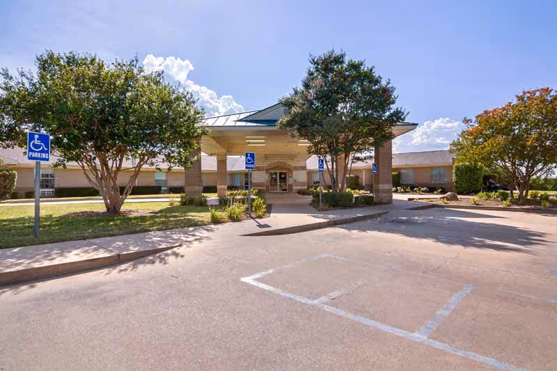 Covered front entrance of a nursing facility with a porte-cochère, accessible parking signs, and trees.