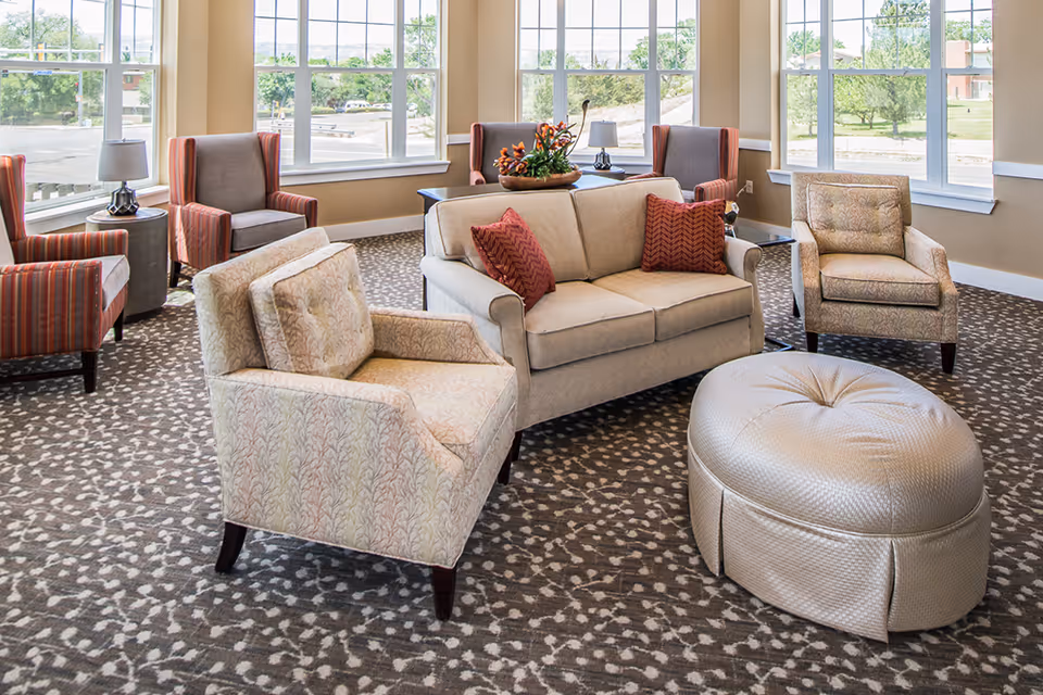 A bright and spacious living room area with large windows letting in natural light. The room features a beige loveseat with two red patterned pillows, two beige armchairs with subtle patterns, two striped armchairs, a round beige ottoman, and side tables with lamps. A decorative flower arrangement is placed on a table behind the loveseat.