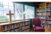 A cozy reading nook in a library with wooden bookshelves filled with books, a large window letting in natural light, a wooden cross on the windowsill, a table lamp, and a comfortable purple armchair.