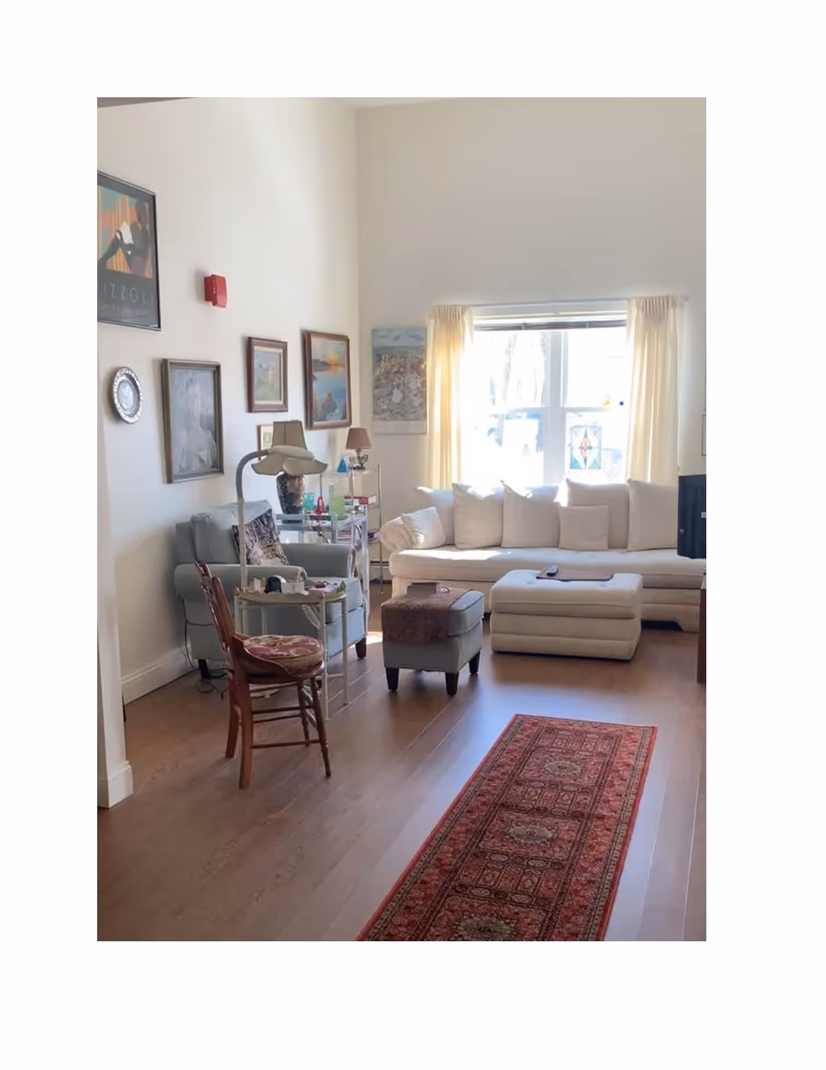 A bright living room with a large window covered by light curtains letting in natural light. The room features a white sectional sofa with multiple cushions, a small ottoman, a light blue armchair, a wooden chair with a patterned cushion, and a red patterned rug on the wooden floor. Several framed paintings and a decorative plate hang on the white walls.