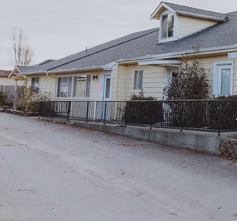 Front exterior of a single-story yellow assisted living building with a driveway, metal railing, and shrubs.