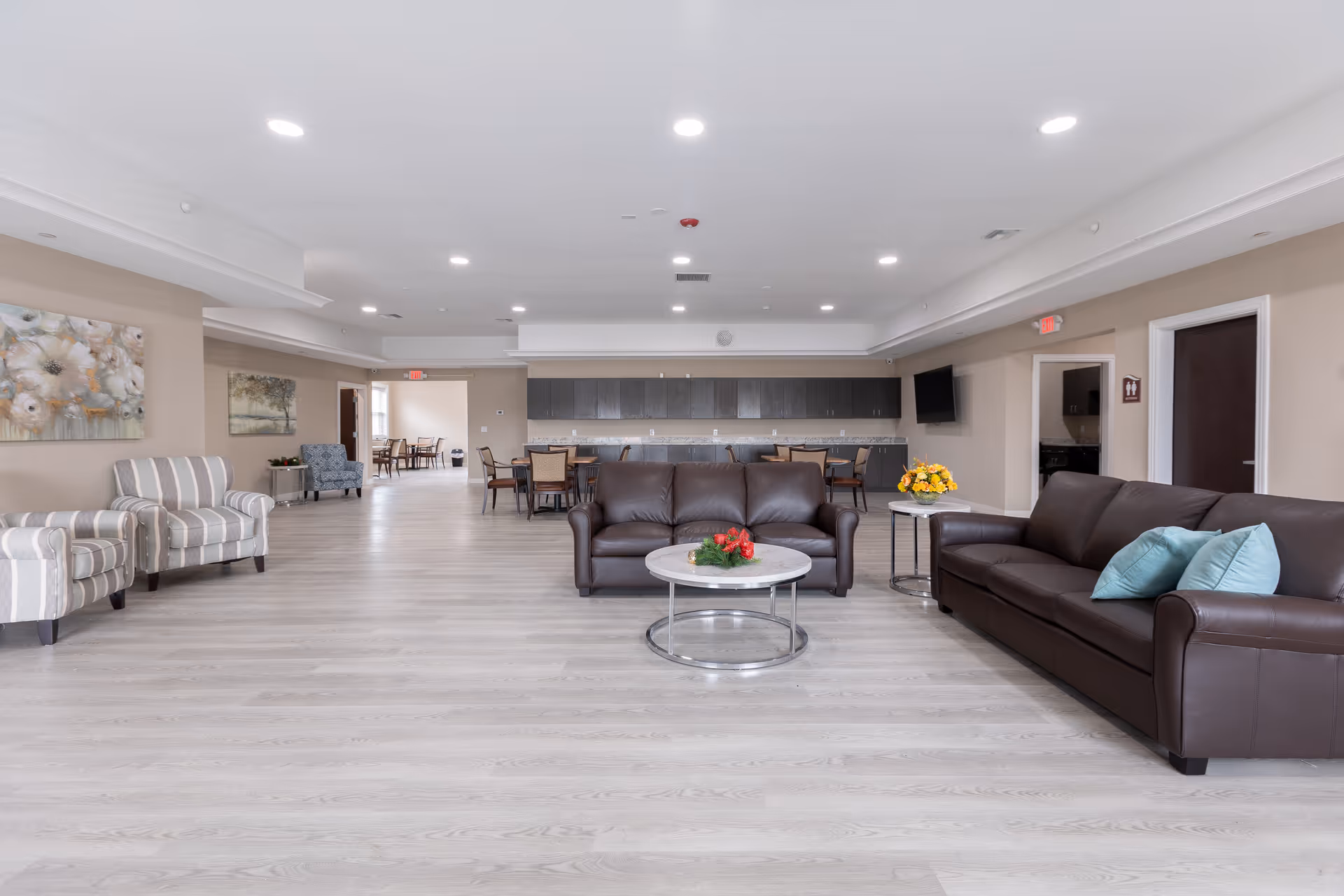 Spacious common lounge in an assisted living facility with leather sofas, armchairs, coffee tables and a dining area with a kitchenette in the background.