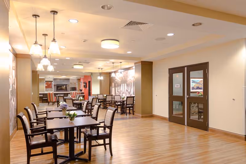 A spacious dining area with multiple tables and chairs arranged neatly. The room features wooden flooring, pendant lights hanging from the ceiling, and a wall with a large decorative mural. Double doors are visible on the right side of the room.