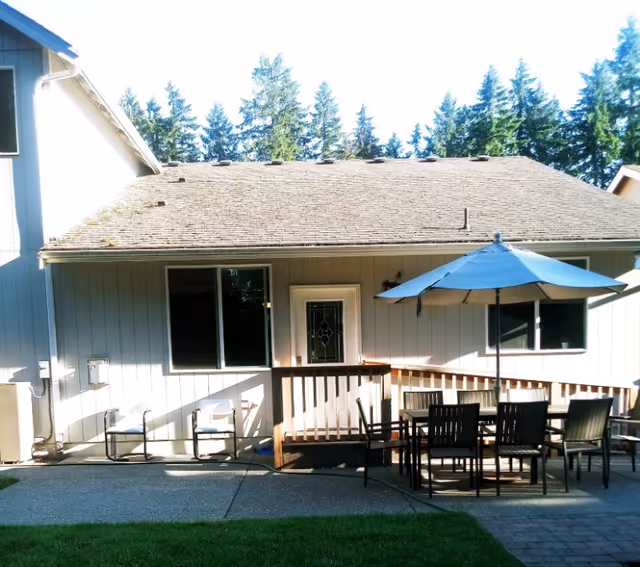 Outdoor patio area of a senior living facility with a table and several chairs under a large blue umbrella, two white chairs along the wall of the building, and a door and windows on the building's exterior. Tall trees are visible in the background.