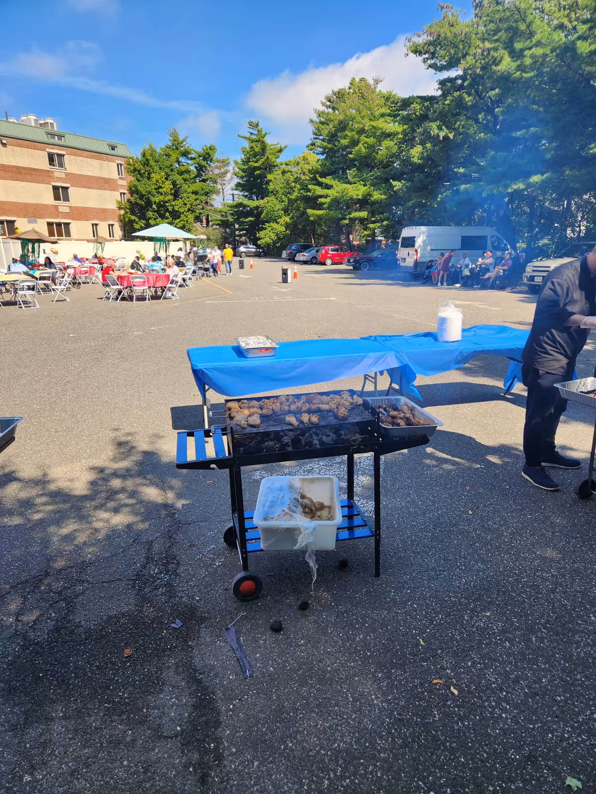 Outdoor barbecue event in a parking lot with a grill cooking meat, a long table covered with a blue tablecloth, and people sitting at tables and standing in the background near a building and trees.