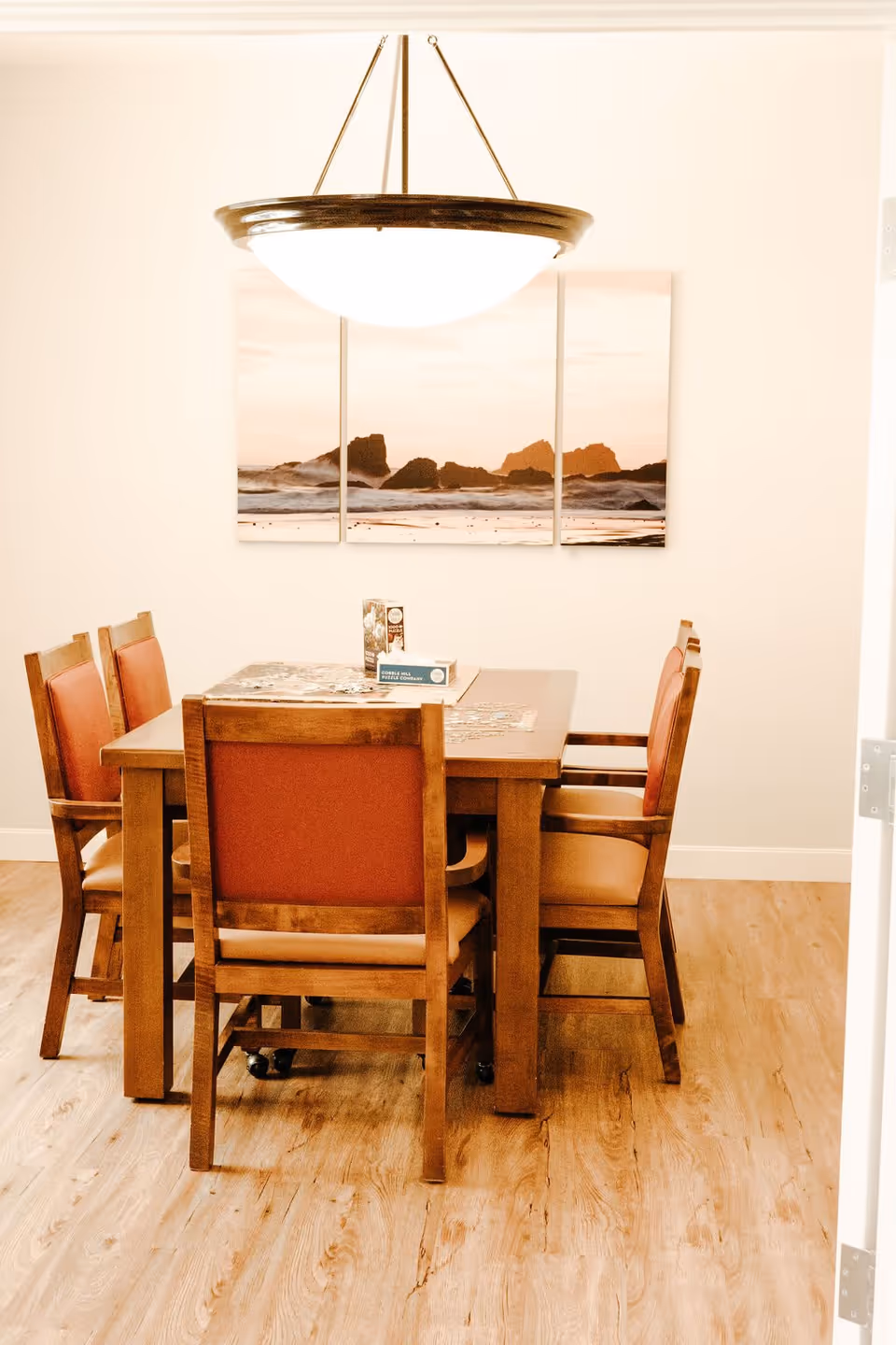 A dining area with a wooden table and six chairs with orange cushions. Above the table hangs a round ceiling light fixture. On the wall behind the table is a three-panel artwork depicting a rocky shoreline at sunset. The floor is wooden.